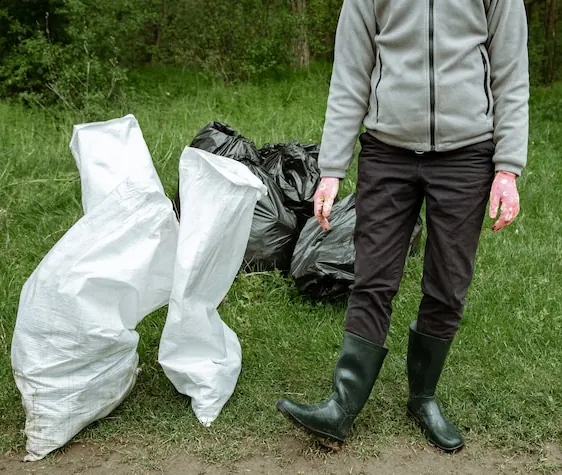 Saci albi de rafie și saci negri cu gunoi pe iarbă, lângă un om în cizme de cauciuc
